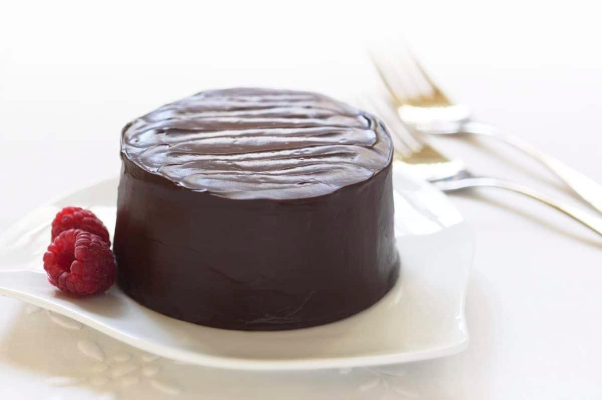 A glossy chocolate cake adorned with fresh raspberries on a white plate.
